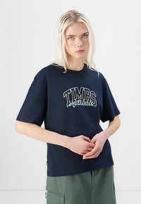 Tamsiai mėlynas medvilninis marškinėliai trumpomis rankovėmis, su balta ir šviesiai mėlyna "TIMBS Authentic" grafika ant priekio. Paprastas dizainas, laisvas siluetas.