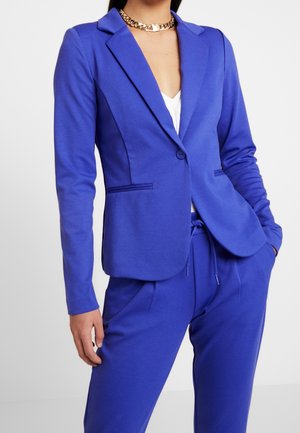 Blau taillierter Blazer und passende Hose, getragen über einem weißen Top, mit einer auffälligen goldenen Kettenkette, vor einem schlichten hellen Hintergrund.