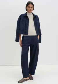 Woman wearing a dark blue denim jacket draped over shoulders, matching wide-leg pants, cream knit sweater, and brown slip-on shoes.