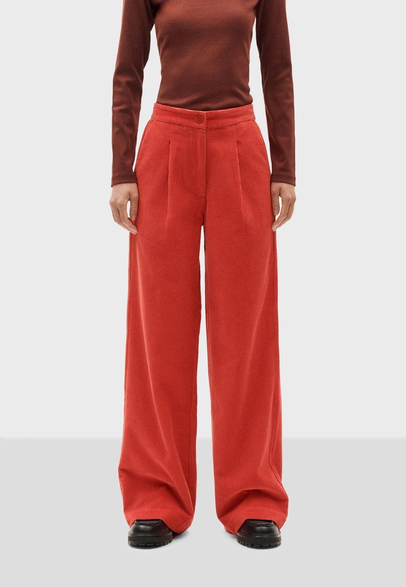 Pantaloni a gamba larga in corda rossa con pieghe frontali, vita alta e chiusura con un bottone. Abbinati a una maglietta marrone a maniche lunghe e scarpe nere.