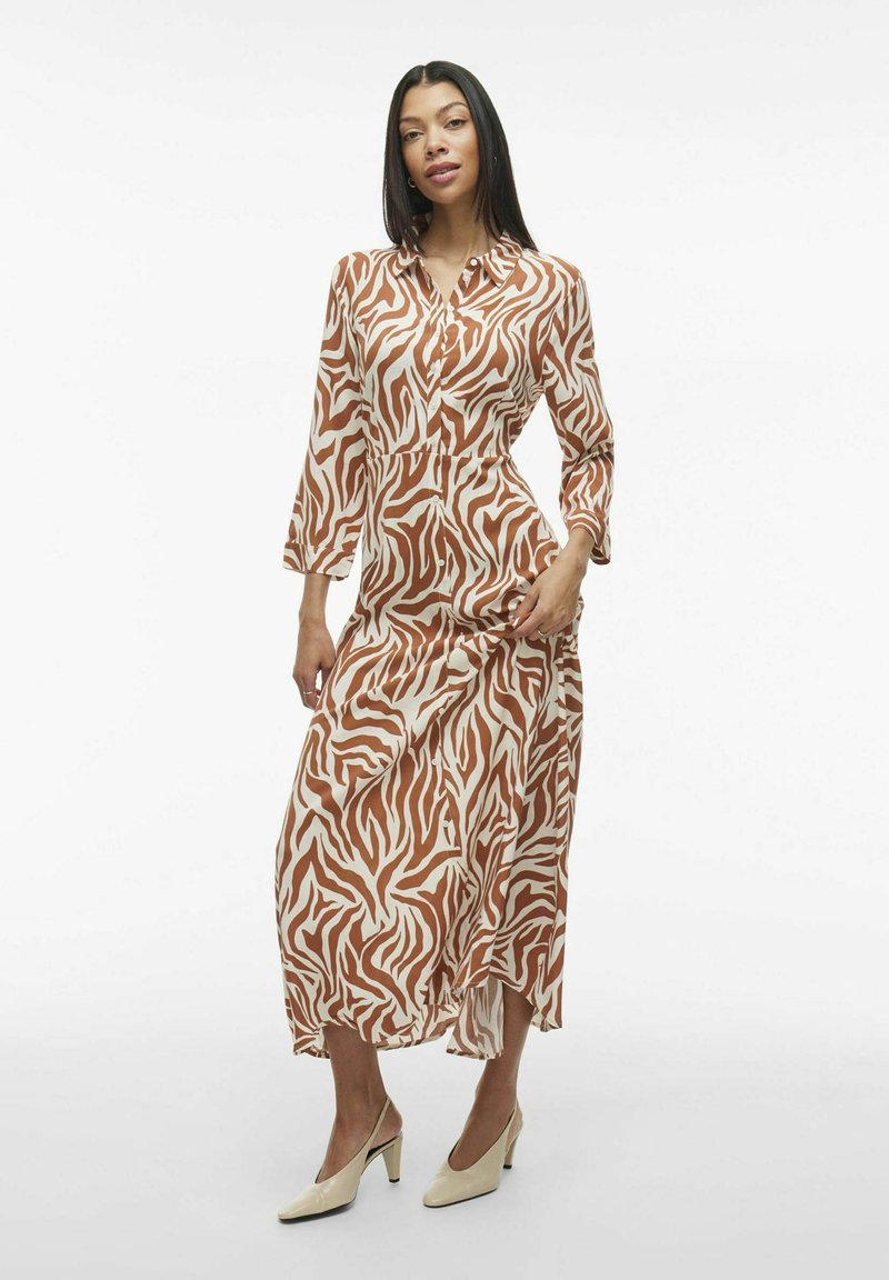 Robe en imprimé zèbre marron et blanc avec des manches longues, des boutons et une silhouette fluide, associée à des chaussures à talons beiges.