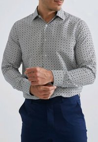 Homme portant une chemise à manches longues grise à motifs, boutonnée au niveau des poignets, associée à un pantalon bleu foncé, ajustant un poignet avec les deux mains.