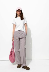 T-shirt blanc à manches courtes, pantalon large rayé, casquette rose clair et sac à bandoulière rose ; sabots bruns avec des accents métalliques. Fond simple.