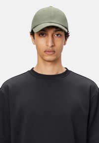 Gorra caqui con una textura suave y diseño de seis paneles, llevada con una sudadera negra de corte holgado. Apariencia minimalista y simplificada.