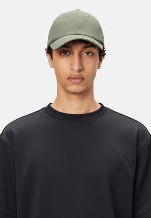 Gorra caqui con una textura suave y diseño de seis paneles, llevada con una sudadera negra de corte holgado. Apariencia minimalista y simplificada.