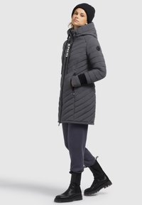 Grå quiltad jacka med hög krage, dragkedjestängning och logodetaljer. Matchad med mörka byxor och svarta ankelstövletter.