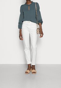 Femme portant un blouse à manches longues teal, un jean taille haute blanc, des sandales à talons en laine beiges, et portant un sac à main en motif serpent.