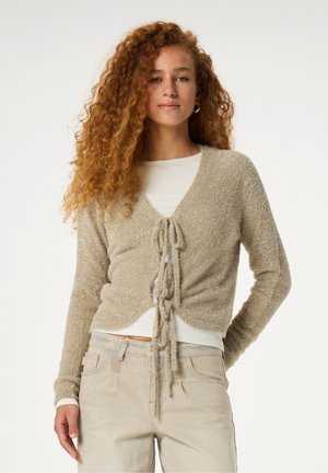 Beige fluffy cardigan with a tie-front closure, featuring a textured finish and long sleeves, worn over a white ribbed top.