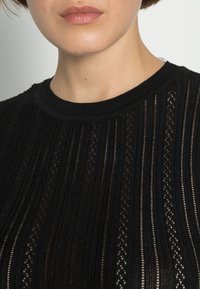 Musta ribbineulottu neulepaita, jossa pystysuorat raidat ja avoimet silmukat, pyöreä pääntie ja sileä pinta.