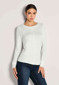Frau mit dunklen Haaren in einem weißen, langärmligen, enganliegenden Oberteil und dunkelblauen Jeans, die vor einem schlichten, hellen Hintergrund steht.