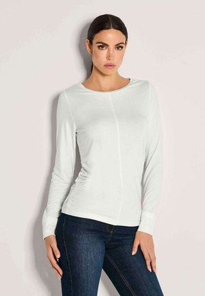Frau mit dunklen Haaren in einem weißen, langärmligen, enganliegenden Oberteil und dunkelblauen Jeans, die vor einem schlichten, hellen Hintergrund steht.
