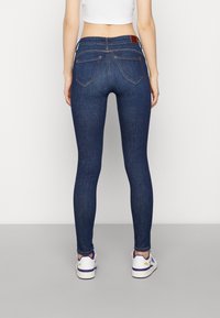 Skinny dark blue jeans with a smooth texture, featuring back pockets and a brown leather patch on the waistband, paired with white sneakers.