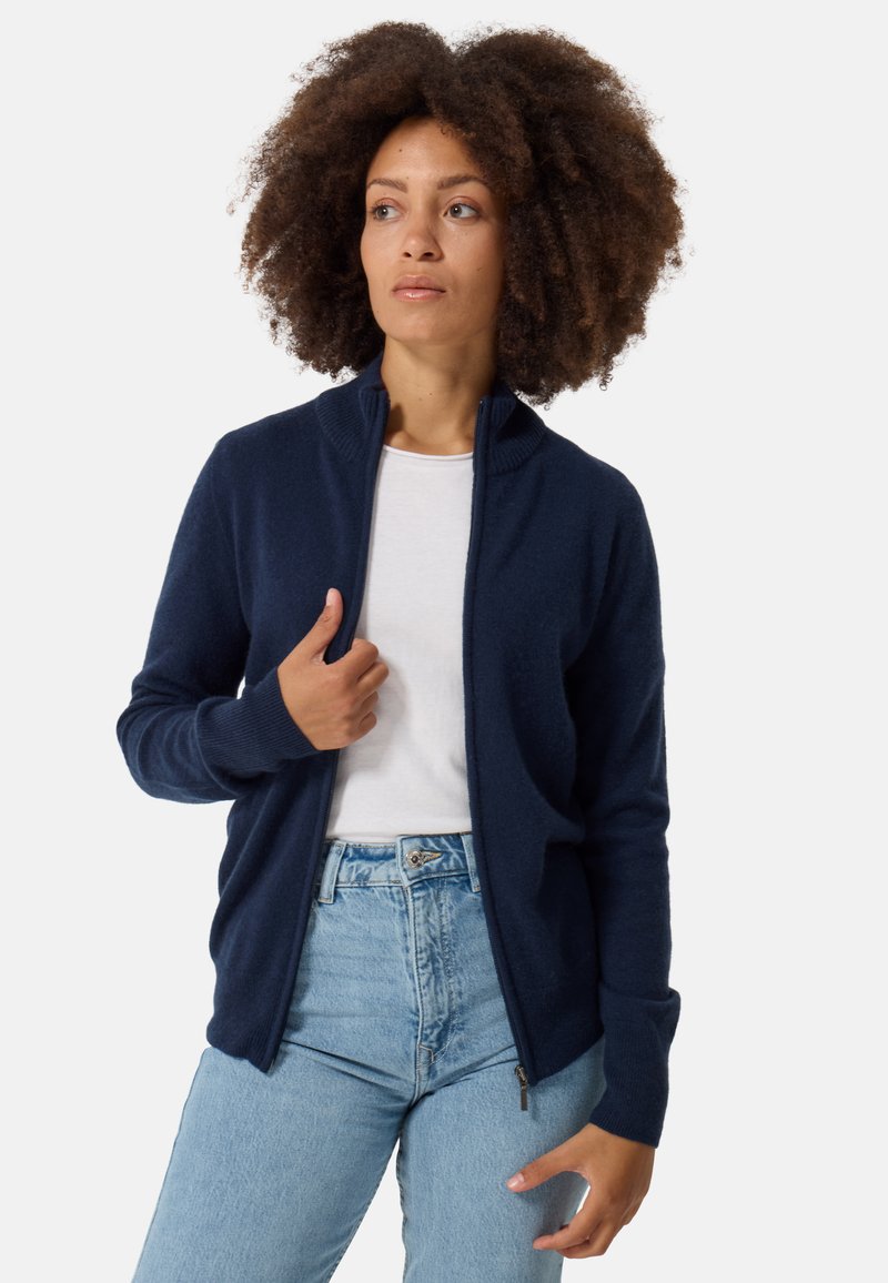 Marineblauer Reißverschlusspullover mit hohem Kragen, weichem Material, langen Ärmeln und gerippten Bündchen. Kombiniert mit einem weißen T-Shirt und hellblauen Jeans.