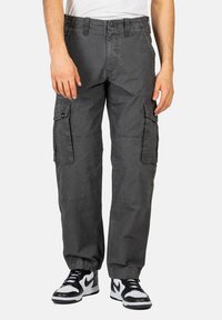 Graue Cargo-Hosen aus Baumwolle, mit mehreren Seitentaschen, einer lockeren Passform und elastischen Bündchen an den Knöcheln. Getragen mit schwarz-weißen Sneakers.