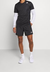 Camiseta atlética negra de mangas cortas con mangas largas blancas por debajo, combinada con pantalones cortos negros y zapatillas blancas. Diseño minimalista.