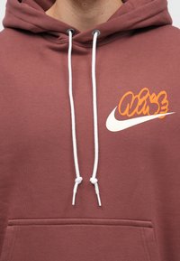 Bordový hoodie s bielou šnúrkou a prednou kapsou. Obsahuje farebný grafický dizajn na hrudi, ktorý kombinuje oranžové a biele prvky.