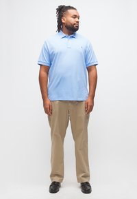 Άνδρας με dreadlocks που φοράει ανοιχτό μπλε πόλο πουκάμισο, μπεζ παντελόνι και μαύρα παπούτσια, στέκεται και κοιτά προς τα δεξιά του.