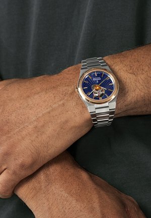 Montre en acier inoxydable argentée avec un cadran bleu à motifs, des accents en or rose et un design à cœur ouvert, portée au poignet sur un fond sombre.