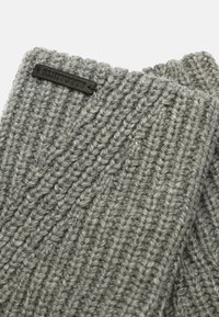 Sciarpa grigia lavorata a maglia con una texture a coste, dotata di una piccola etichetta nera rettangolare con testo in rilievo. Materiale morbido e caldo.