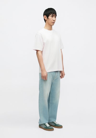 Ανοιχτό ροζ βαμβακερό μπλουζάκι με κοντά μανίκια, συνδυασμένο με ανοιχτό μπλε χαλαρά τζιν και πράσινα αθλητικά παπούτσια με λευκά κορδόνια και καφέ σόλες.