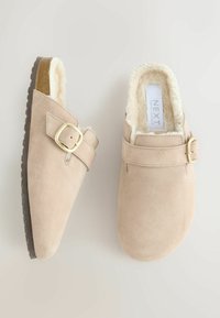 Zuecos de gamuza beige con un suave forro de piel sintética blanca, que presentan un detalle de hebilla dorada y una suela de goma texturizada marrón.