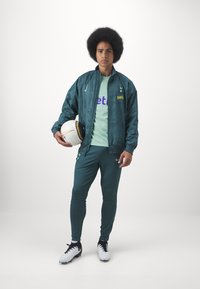 Tuta verde con giacca a fantasia, camicia verde chiaro con logo, pantaloni verde scuro e scarpe da calcio nere; tiene in mano un pallone da calcio bianco.