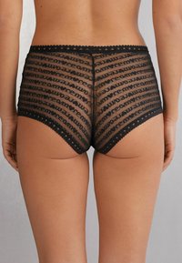 Zwarte mesh slipjes met een herhalend tekstpatroon. Voorzien van een geschulpt kantdetail en een soepele, rekbare tailleband. Hoog uitgesneden, volwaardige dekking.