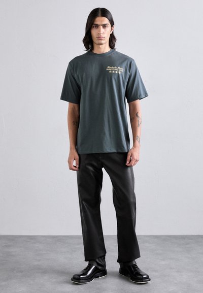 Camiseta de algodón verde oscuro con mangas cortas y cuello redondo, con un logotipo gráfico dorado en el pecho. Combinada con pantalones y zapatos negros.