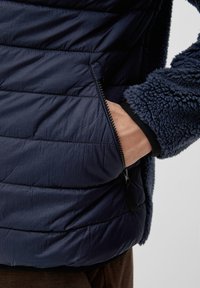 Giubbotto puffer blu navy con design imbottito, dotato di tasca laterale con zip e maniche in morbido e testurizzato fleece. Dettaglio con polsini elasticizzati incluso.