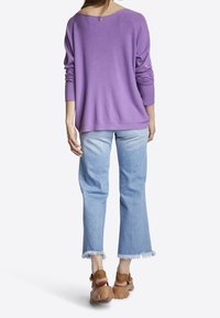 Fioletowy, oversized sweter z szerokim dekoltem, w połączeniu z jasnoniebieskimi dżinsami w stylu dzwonów z postrzępionymi brzegami i brązowymi, grubymi sandałami.