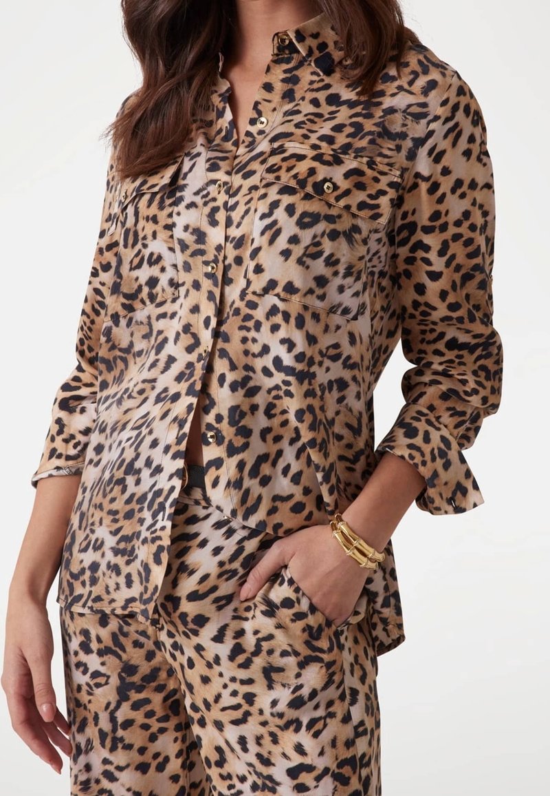 Femme portant une chemise et un pantalon assortis à imprimé léopard, une main dans la poche, bracelet doré au poignet, cheveux lâchés, fond uni.