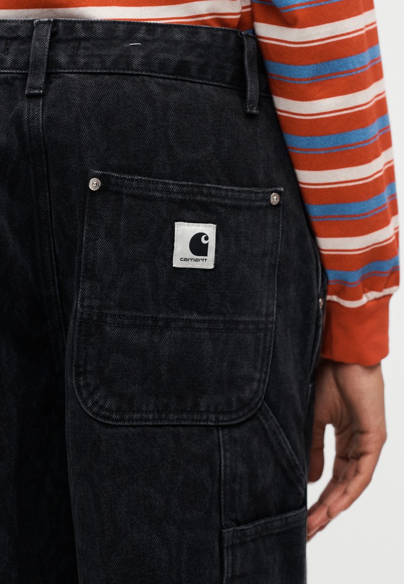 Person trägt schwarze Carhartt-Hose mit großer Gesäßtasche und ein orange-blau-weiß gestreiftes Langarmshirt.