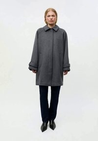 Casaco de lã cinza com gola, fecho de botão e punhos largos. O modelo veste calças escuras e sapatos pretos pontiagudos. Design minimalista, sem padrões.