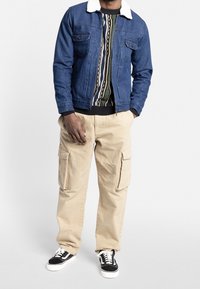 Veste en jean avec col en fausse fourrure, intérieur en tricot à motifs, pantalon cargo kaki et sneakers noirs avec accents et lacets blancs.