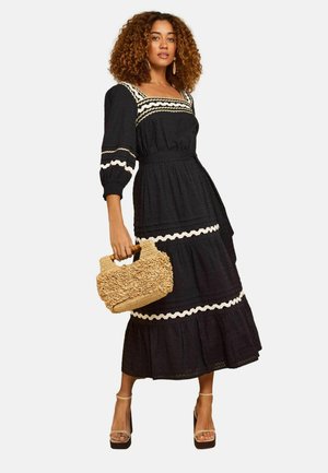 Sieviete ar cirtainiem matiem, valkājot melnu midi kleitu ar baltu apdari, turot austu somu un nēsājot bēša krāsas platformu sandales.