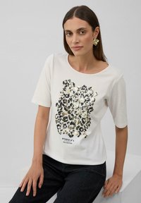 Biała bawełniana koszulka z krótkim rękawem, ozdobiona błyszczącym złotym i czarnym wzorem w panterkę oraz napisem "STUDIO N°1 COLLECTION" na dole.