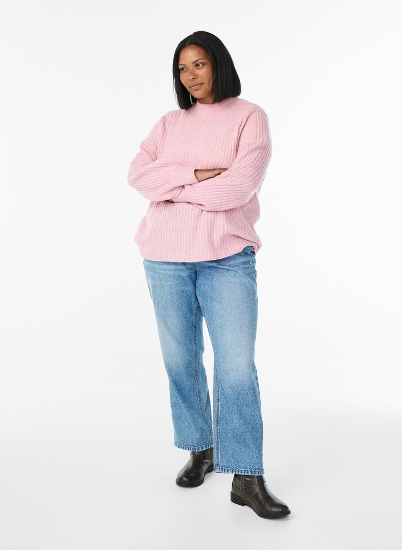Frau mit mittellangen schwarzen Haaren, die einen pinkfarbenen Pullover, blaue Jeans und schwarze Stiefeletten trägt, steht mit verschränkten Armen da.