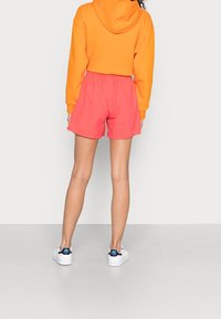 Eine Person, die einen leuchtend orangefarbenen Hoodie, korallenrosa Shorts und weiße Sneakers trägt, steht mit dem Rücken zu einem einfarbigen hellen Hintergrund.