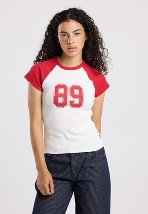 T-shirt en coton avec des manches raglan rouges, un corps blanc et un grand "89" rouge imprimé sur le devant. Manches courtes et design ajusté.