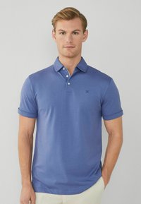Uomo biondo che indossa una polo blu a maniche corte con un piccolo logo ricamato sul petto, in piedi davanti a uno sfondo grigio uniforme.