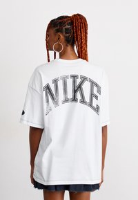 Vrouw met lange rode vlechten, die grote gouden oorbellen draagt en een wit oversized Nike T-shirt met zwarte letters aan heeft, met haar gezicht weggedraaid van de camera.