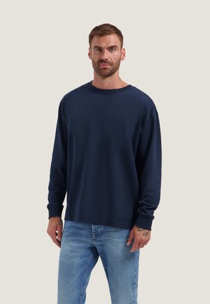 Uomo in piedi che indossa una maglia a maniche lunghe blu navy con scollo tondo e jeans azzurri su uno sfondo chiaro uniforme.