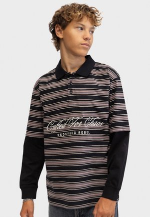 Jeune personne aux cheveux bouclés portant un polo rayé par-dessus un t-shirt noir à manches longues, regardant sur le côté sur un fond uni.
