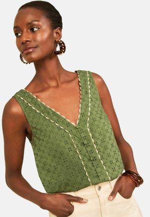 Femme portant un chemisier sans manches vert olive à œillets avec une bordure festonnée et un pantalon beige, accessoirisée de grandes boucles d’oreilles créoles et de bracelets.
