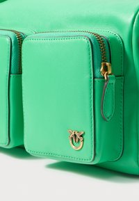 Pocket di una borsa in pelle verde con zip dorata e piccolo emblema dorato che raffigura due uccelli su un anello.