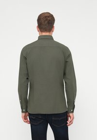 Camisa verde de manga larga hecha de algodón con cuello estructurado, puños abotonados y corte recto, llevada con jeans de mezclilla oscura.