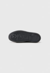 Semelle de sneaker noire avec des rainures texturées et des accents en caoutchouc circulaires. Présente un logo en écriture sur la surface, conçu pour l'adhérence.