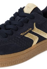 Gros plan d'une sneaker noire avec dessus texturé, semelle épaisse marron, bande latérale dorée et logo "Tamaris" près des lacets noirs et de la languette beige.