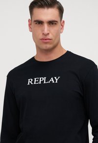 Schwarzes Langarm-T-Shirt aus Baumwolle mit einem weißen "REPLAY"-Logo auf der Brust, Rundhalsausschnitt und schlanker Passform.
