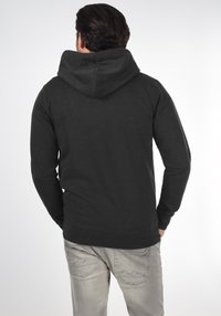 Mand vender væk iført en sort hættetrøje og lysegrå jeans mod en ensfarvet hvid baggrund.